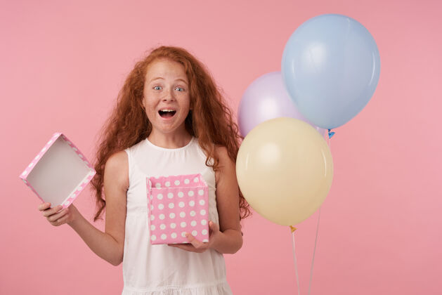 童年摄影棚拍摄的可爱红发女孩手持礼品包装盒 脸上洋溢着兴奋的神情 扬起眉毛 快乐地看着镜头 站在粉色背景下 手里拿着彩色气球嘴欧洲举行