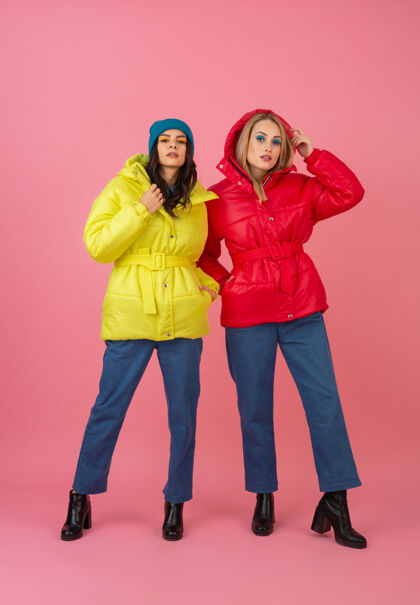 年轻两个迷人的女孩在粉红色的背景下 穿着鲜艳的红色和黄色的彩色冬季羽绒服摆姿势女士女孩时尚