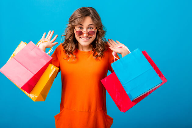 蓝色图片中的兴奋迷人的微笑时尚女性购物狂穿着橙色的新潮连衣裙拿着购物袋在蓝色的工作室背景隔离商店包持有