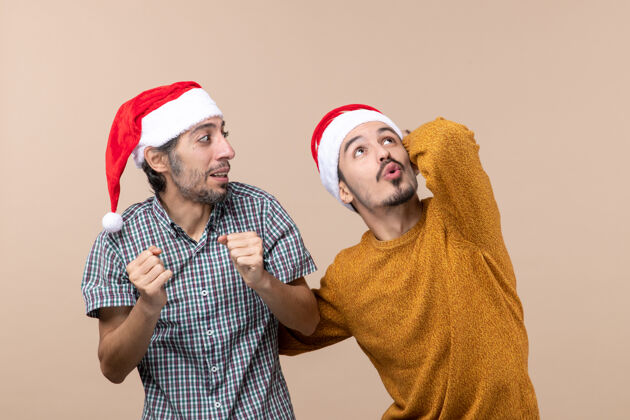 米色正面图两个戴着圣诞帽的家伙 一个在米色孤立背景下惊讶地看着高处男人伙计们视图