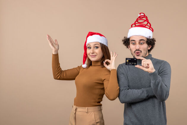 红色圣诞节心情激动满意酷情侣戴着红色圣诞老人帽展示银行卡 女人做ok手势穿着心情快乐