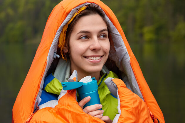 独自微笑积极的年轻女旅客裹着睡袋 在寒冷的秋日里用热饮取暖业余时间人类积极