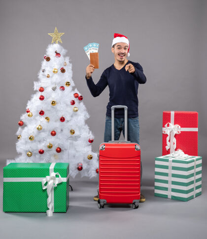 购物兴奋的年轻人拿着手提箱在圣诞树周围展示他的旅行票和灰色的礼物帽子年轻圣诞帽
