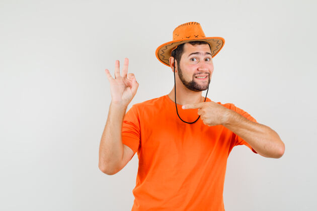 休闲年轻人指着自己的ok标志 穿着橙色t恤 戴着帽子 看上去很高兴 正对着前方时尚表情帽子