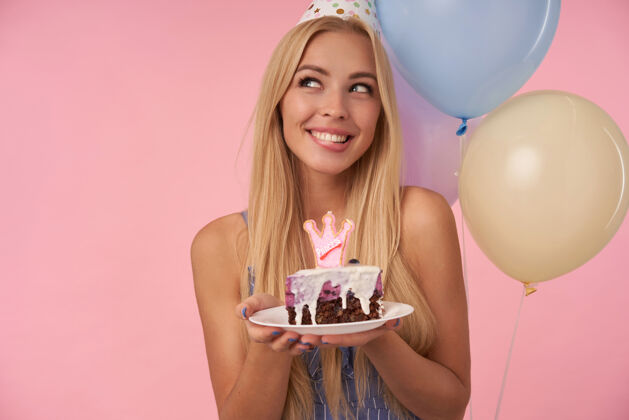裙子迷人的金发女郎 手里拿着一块蛋糕 面带幸福的笑容 神魂颠倒地看着一边 穿着蓝色的夏装 戴着圆锥形的帽子 站在粉色的背景上微笑帽子美丽
