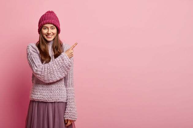 指向乐观的欧洲女性指着右上角 穿着时髦的冬装 温柔地微笑 做广告 隔着粉色的墙看促销的空白积极毛衣空白空间