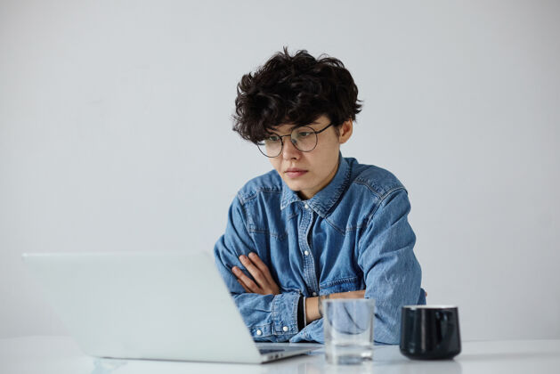 茶一张室内照片 年轻的黑发卷发女子戴着眼镜 手持笔记本电脑 穿着蓝色牛仔裤衬衫 坐在白色背景上 在现代办公室工作时尚自然发型