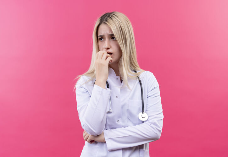 医学悲伤的医生年轻的金发女孩穿着医用长袍戴着听诊器把她的手放在下巴上孤立的粉红色背景金发穿着听诊器
