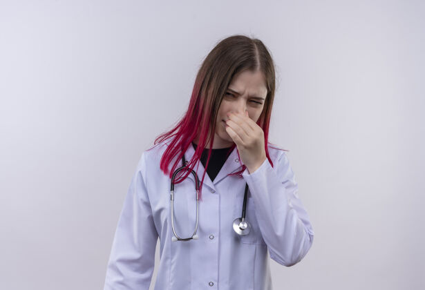穿着穿着听诊器医用长袍的神经质的年轻女医生在孤立的白色背景下闭着鼻子医生女孩鼻子