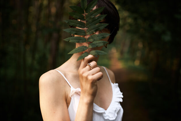 浪漫穿着白色束带裙的神秘少女在森林里独自摆姿势 脸上覆盖着巨大的蕨类植物叶子黑发华丽植物