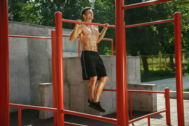 男性在阳光明媚的夏日 一个年轻的白种人在操场上做单杠引体向上在户外训练他的上半身运动 锻炼 健康的生活方式 幸福的概念胸部健康年轻