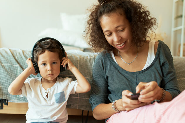 小室内肖像 欢快的西班牙年轻女性手持手机 为她可爱的小儿子播放音乐 小儿子通过无线耳机听歌放松女人可爱