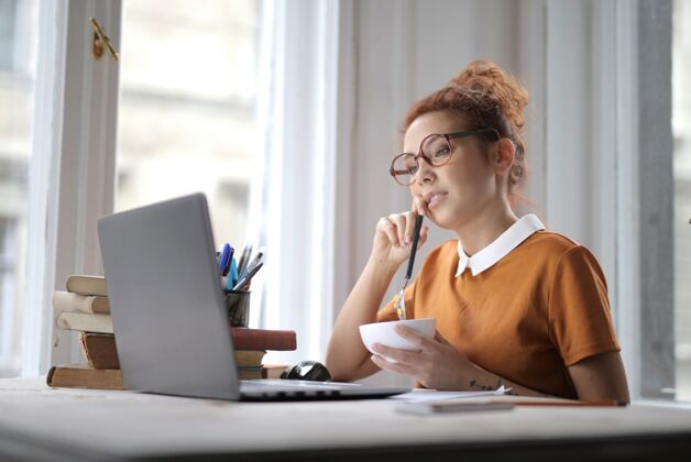 休闲迷人的女性 戴着眼镜 手里拿着一碗麦片粥 坐在桌上的笔记本电脑前电脑女人成人