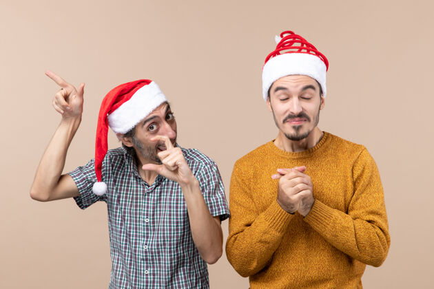 伙计们正面图两个戴着圣诞帽的快乐的家伙一个展示着什么另一个闭上眼睛 双手合十在米色的孤立背景上男人米色两个快乐的家伙