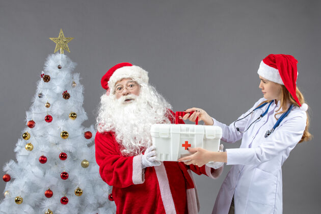 节日圣诞老人和女医生的前视图 女医生在灰色的墙上拿着急救箱圣诞快乐圣诞老人十二月