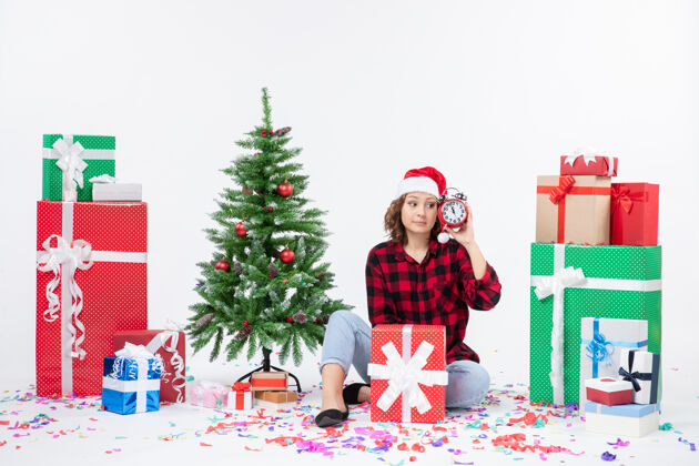 十二月前视图的年轻女子坐在圣诞礼物周围拿着白色墙上的时钟礼物节日前面