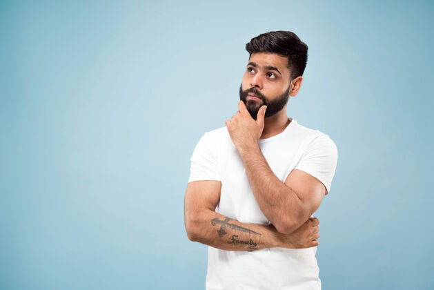 人蓝色背景上穿着白衬衫的印度教年轻人的半身特写肖像人类情感 面部表情 广告概念负面空间手拉胡子思考选择企业家男人工作