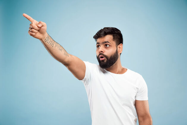 公司蓝色背景上穿着白衬衫的印度教年轻人的半身特写肖像人类情感 面部表情 销售 广告概念负面空间向上指震惊和惊讶成功高管工作