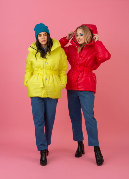 女士两个迷人的女孩在粉红色的背景下 穿着鲜艳的红色和黄色的彩色冬季羽绒服摆姿势衣服蓝色休闲