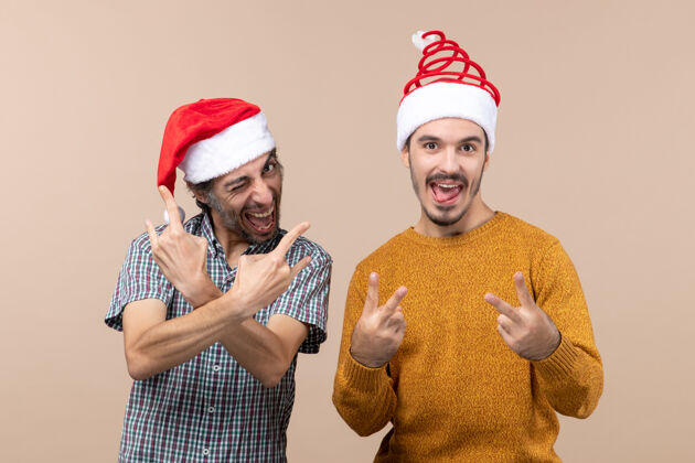 伙计们正面图两个戴着圣诞帽的快乐的家伙一个在制作摇滚标志 另一个在米色孤立背景上的胜利和平标志胜利快乐米色