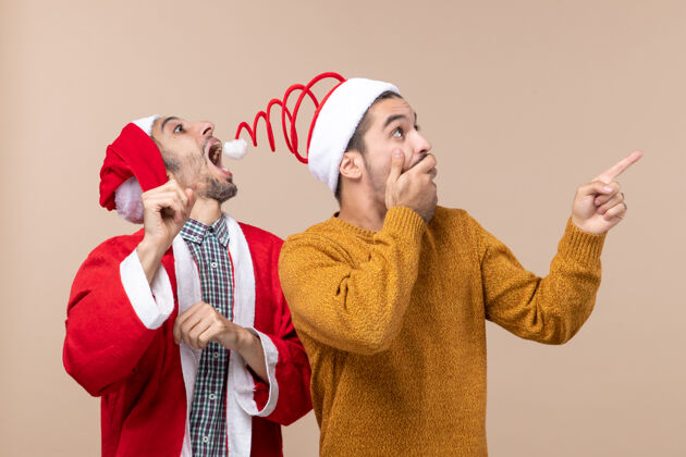 圣诞老人前视图两个朋友与圣诞帽一个试图抓住帽子蓬蓬和其他显示方向的米色孤立的背景视图两个男人