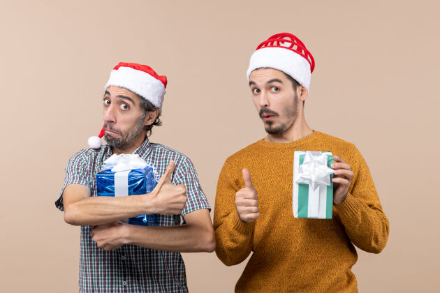 前面正面图两个满意的家伙做拇指向上的标志 并举行在米色孤立的背景圣诞礼物男性向上视图
