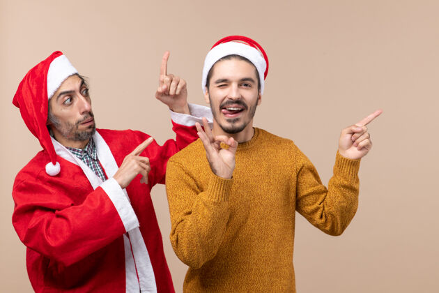 成人前视图两个戴着圣诞帽的朋友都展示了米色背景的东西两者两个视图