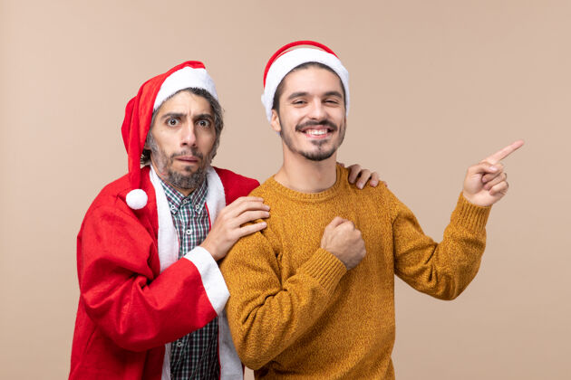 展示正面图两个戴着圣诞帽的朋友 一个抱着朋友的肩膀 另一个在米色孤立的背景上显示方向男性视图肩膀
