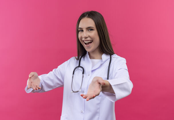 听诊器微笑着的年轻医生女孩穿着听诊器医用长袍假装拿着一个孤立的粉红色背景的东西医生年轻微笑