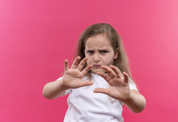 展示悲伤的小女孩穿着白色t恤 在孤立的粉色背景上显示停止手势白色学校T恤