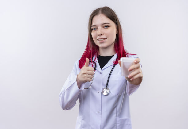 白色高兴的年轻女医生穿着听诊器医用长袍拿着空的大拇指在孤立的白色背景上女孩年轻长袍