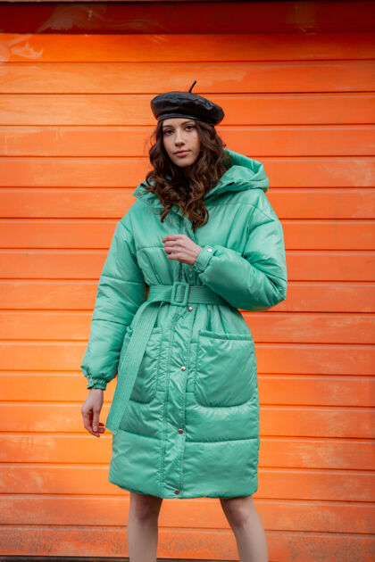 街头穿着冬秋时尚潮流蓬松外套和帽子贝雷帽的时髦女人在街上对着橙色的墙壁摆姿势时尚城市夹克