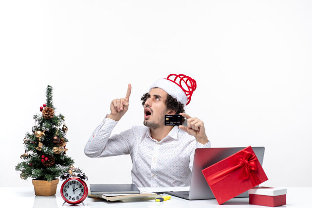 年轻圣诞节心情与年轻的胡须惊讶的商人与圣诞老人帽子持有银行卡 并在办公室指着上面圣诞节持有商务