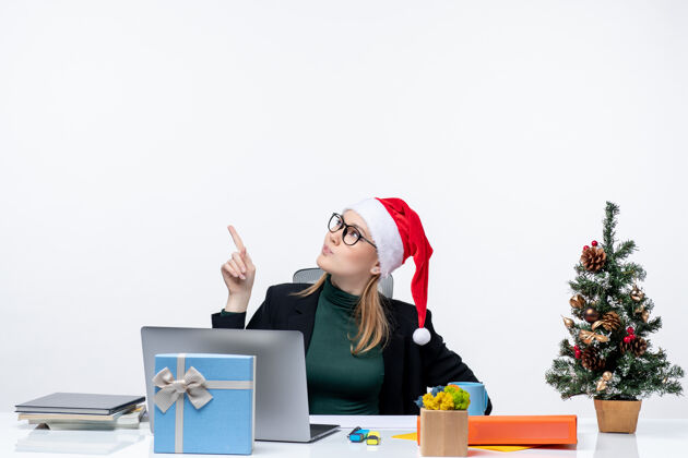 惊讶一位带着圣诞老人帽子的严肃的商务女士坐在一张桌子旁 桌子上有一棵圣诞树 上面有一份礼物 背景是白色的帽子商务圣诞节
