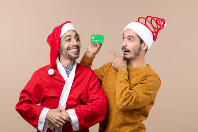 快乐前视图两个快乐的家伙一个穿着圣诞外套 另一个拿着信用卡 在米色孤立的背景下看着它两个快乐的家伙男性微笑
