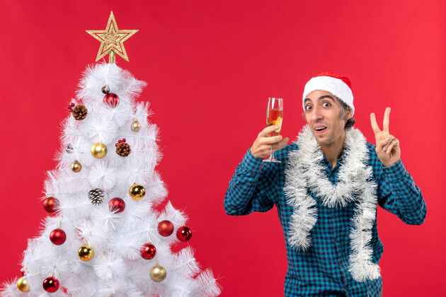 男人圣诞节心情与情绪激动的年轻人圣诞老人帽子在一个蓝色条纹衬衫举行一杯葡萄酒 并显示两个靠近圣诞树圣诞树年轻人帽子