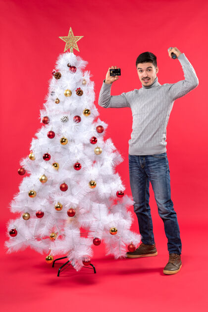 年轻圣诞气氛严肃的年轻人站在装饰过的圣诞树旁 拿着麦克风和电话展示他的力量电话年轻人装饰
