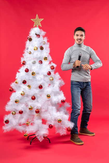自信圣诞节的心情与自信的家伙穿着牛仔裤站在装饰圣诞树附近手持麦克风 愉快地说人男人装饰