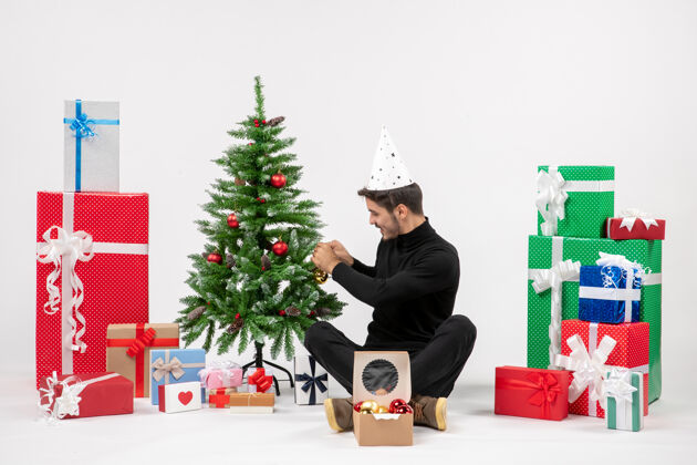 坐着年轻人围坐在节日礼物装饰在白色墙上的小树的前视图圣诞快乐礼物礼物
