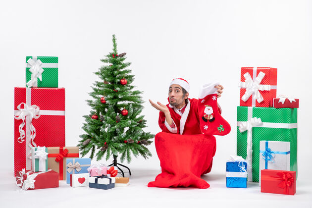 坐着惊喜的圣诞老人正坐在地上 手里拿着圣诞袜 旁边摆放着礼物和装饰在白色背景上的新年树圣诞拿着袜子