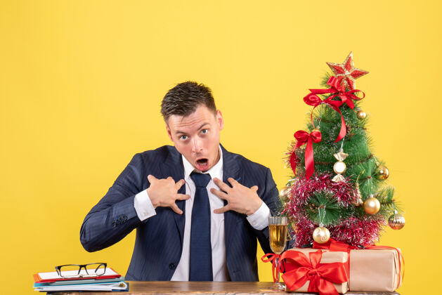 好奇的男人一个好奇的男人把手放在胸前 坐在圣诞树旁的桌子上 用黄色的礼物送给他树礼物桌子