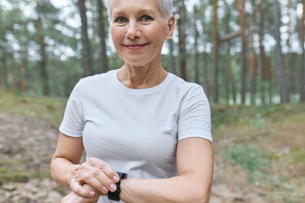 有氧穿着白色t恤的美丽中年妇女的不规则肖像调整手腕上的智能手表运动小工具慢跑者