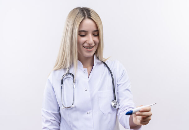 戴微笑的医生年轻的金发女孩戴着听诊器和医用长袍 戴着牙套 在孤立的白色背景上看着手上的温度计年轻女孩医生