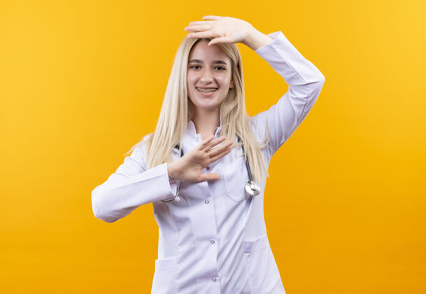 戴微笑的医生年轻的女孩穿着医用长袍戴着听诊器和牙套把她的手放在头上另一个头下孤立的黄色背景黄色微笑长袍