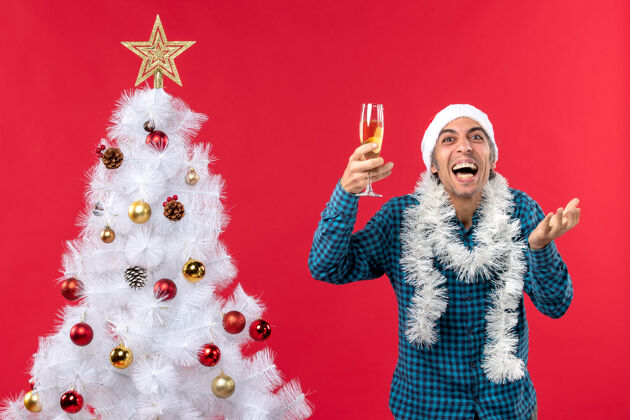 派对圣诞节心情与情绪激动的年轻人圣诞老人帽子在一个蓝色的衬衫剥离举行一杯葡萄酒 并笑着靠近圣诞树年轻人抱着圣诞