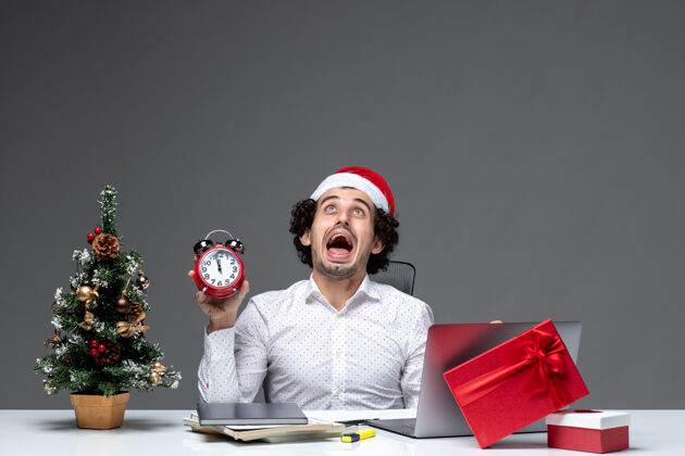 举行年轻愤怒的商人戴着圣诞老人的帽子 拿着钟 坐在黑暗的背景下的办公室里男性成人时钟