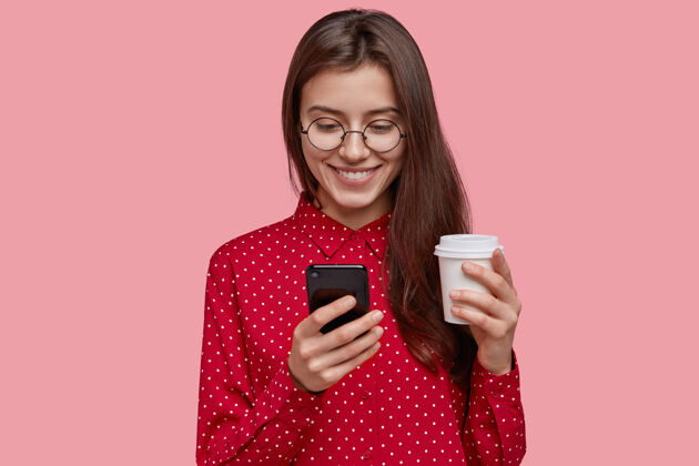 短信妩媚喜庆的年轻女子捧着热腾腾的外卖咖啡 手机 欣然接受了新设备作为礼物 穿着红衬衫 露出了温柔的笑容观看手机手机