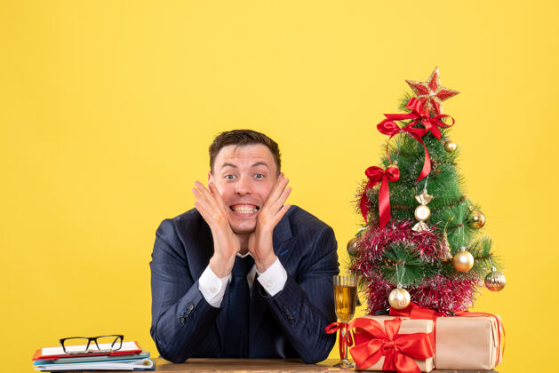 人年轻人在脸颊边放着遮阳帘 坐在圣诞树旁的桌子旁 拿着黄色的礼物黄色礼物商人