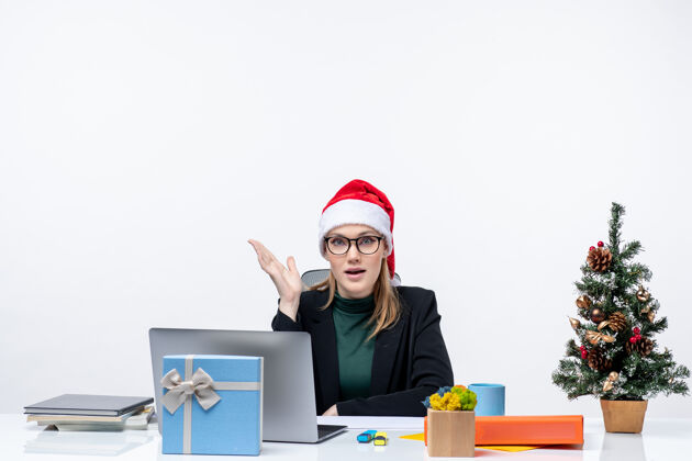 圣诞树办公室里 一位好奇迷人的女士戴着圣诞老人的帽子坐在桌子旁 桌上放着圣诞树和礼物桌子电脑女人