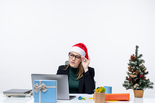 公司专心致志的女商人 戴着圣诞老人帽 坐在一张桌子旁 桌上放着圣诞树和礼物 在白色背景上查看邮件桌子圣诞节办公室